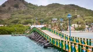 Ponte de madeira pintada de cores vivas sobre um corpo de água até chegar a uma pequena cidade litorânea em San Andres. Ao fundo, uma montanha de vegetação esparsa.