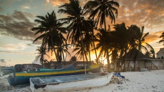 Pôr do sol em San Andres, com o céu em tons quentes de laranja e amarelo, contrastando com as nuvens dispersas. Na areia da praia, há barcos de pesca em repouso.