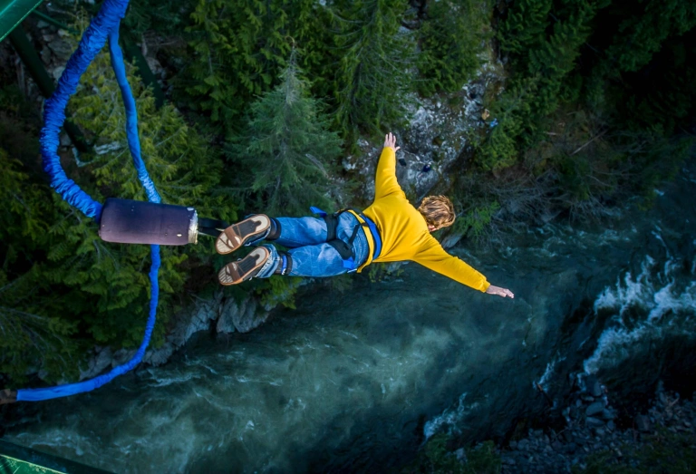 Homem branco e loiro, vestindo uma calça jeans e uma blusa amarela, salta de bungee jump sobre um rio com uma floresta às margens.