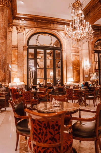 Foto de bar clássico com ricos ornamentos em tons bronzeados em Buenos Aires, Argentina. Destacam-se muitas mesas e cadeiras de madeira maciça, grande lustre e imponente porta de vidro.