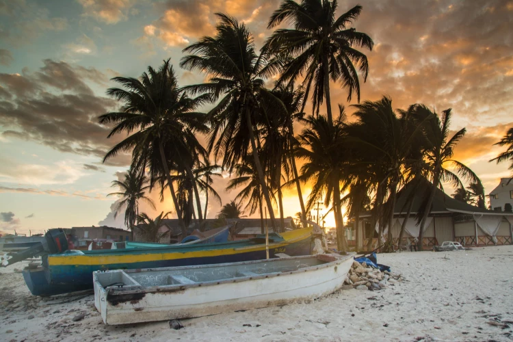 Pôr do sol em San Andres, com o céu em tons quentes de laranja e amarelo, contrastando com as nuvens dispersas. Na areia da praia, há barcos de pesca em repouso.